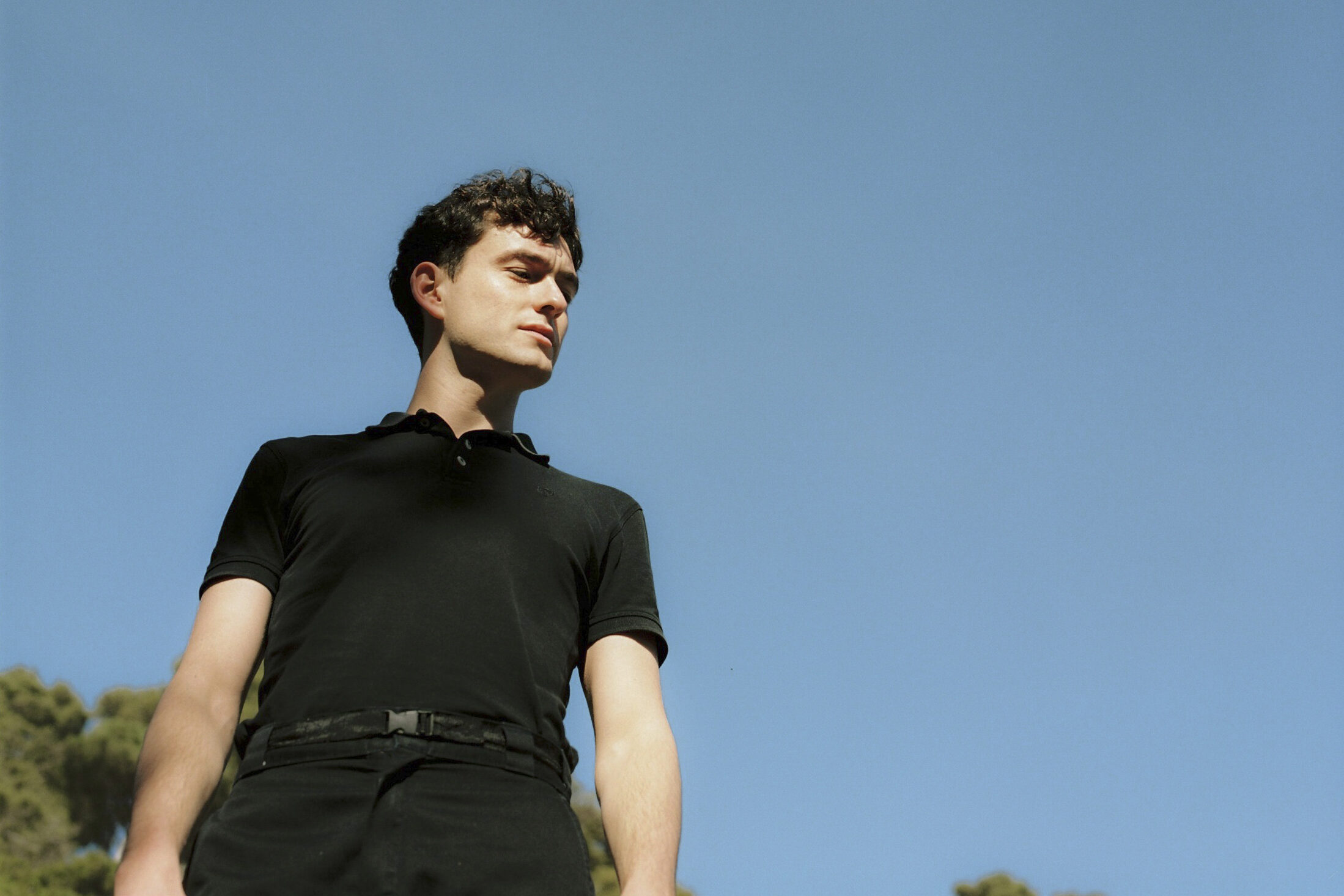 A person wearing a black shirt stands outdoors against a clear blue sky, looking to the side. The perspective is from below, emphasizing the sky and creating a serene atmosphere. Trees are faintly visible in the bottom corners.