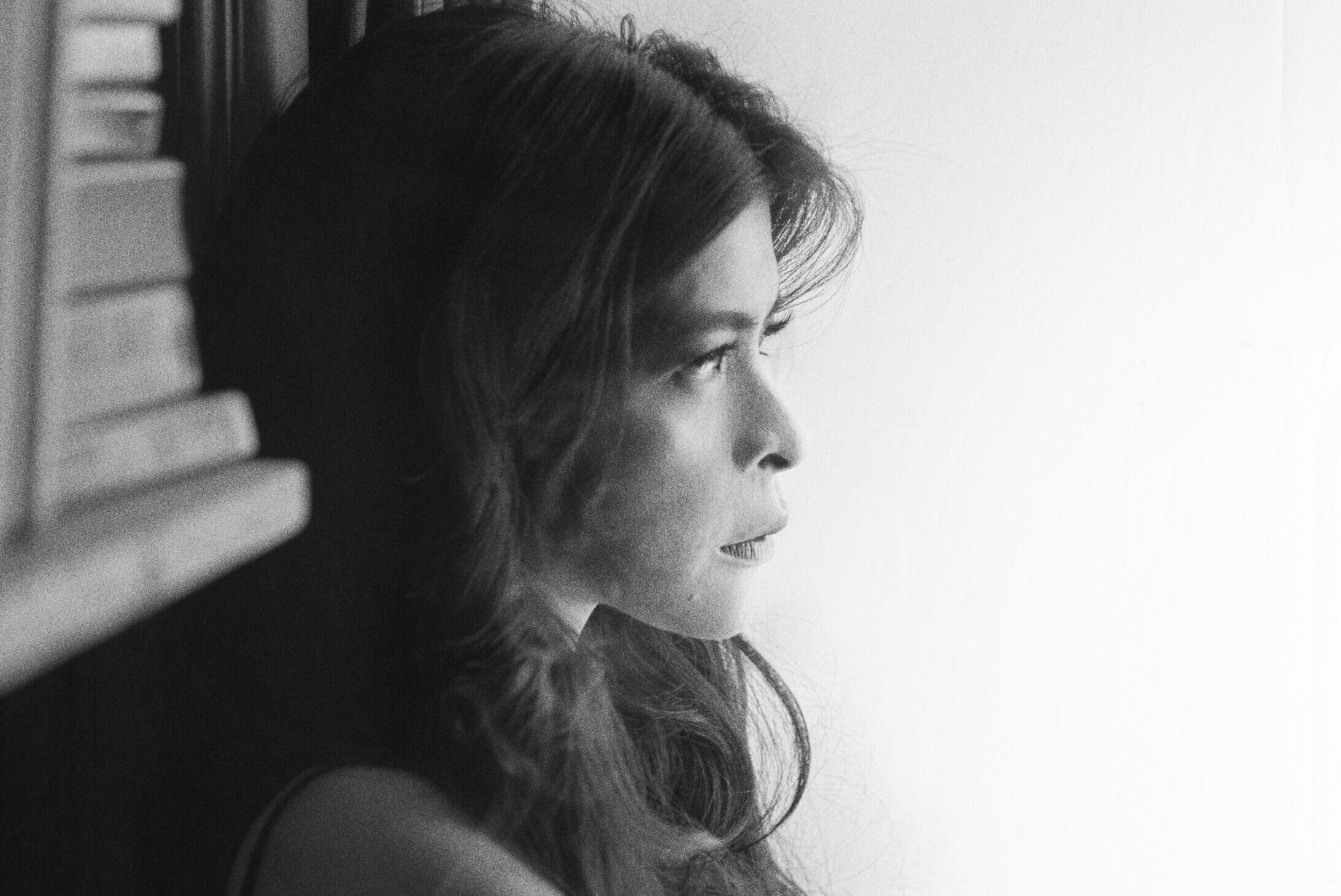 Black and white photo of a person with long hair looking pensively out a window. Theyre holding a cigarette in one hand and are dressed in a sleeveless top. Books are visible on a shelf beside them.