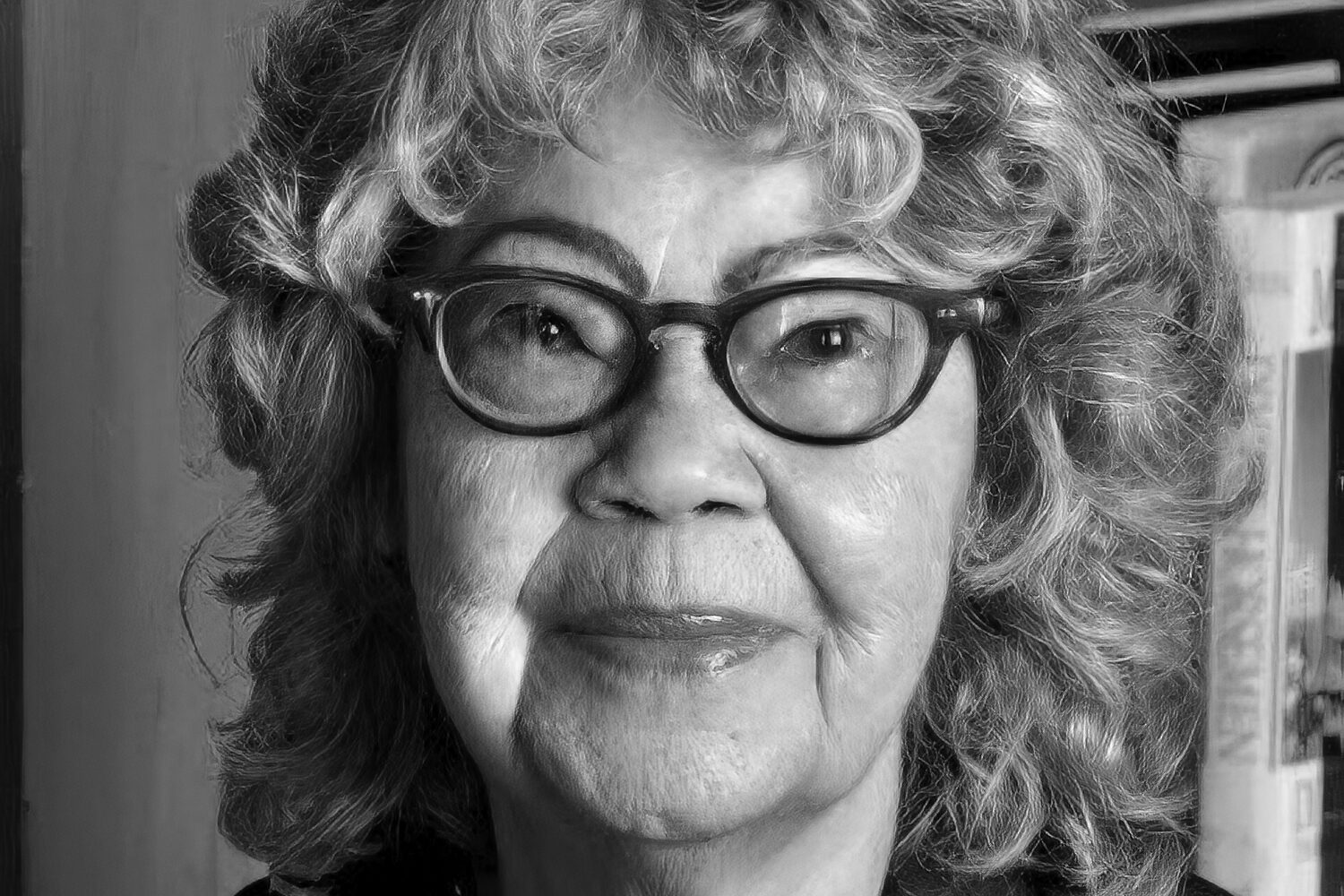 Elderly person with curly hair and glasses, wearing a necklace and a dark top, looking at the camera with a neutral expression in a black-and-white photo.
