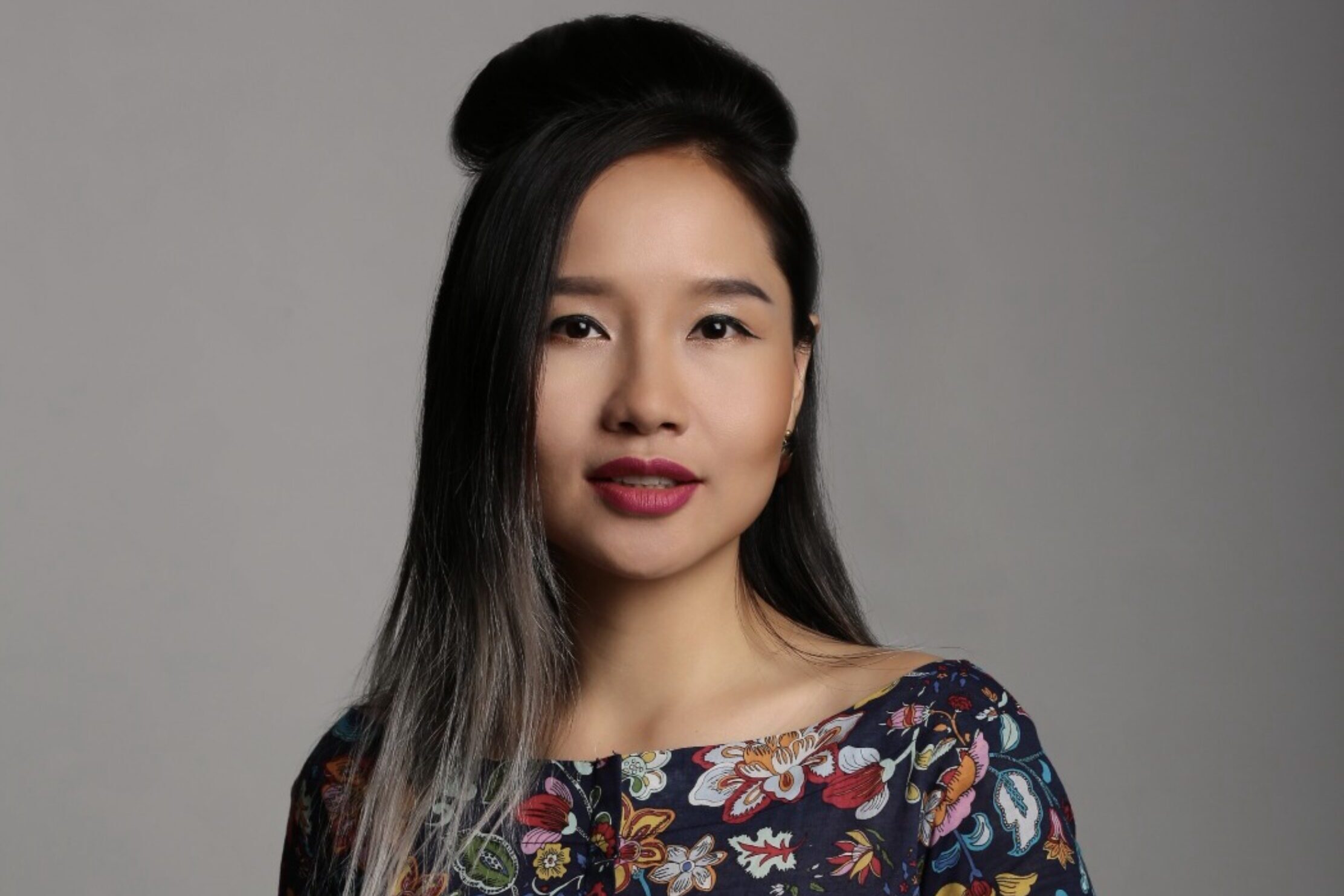 Woman with long, straight hair and an updo, wearing a colorful floral top, looking at the camera against a plain background.