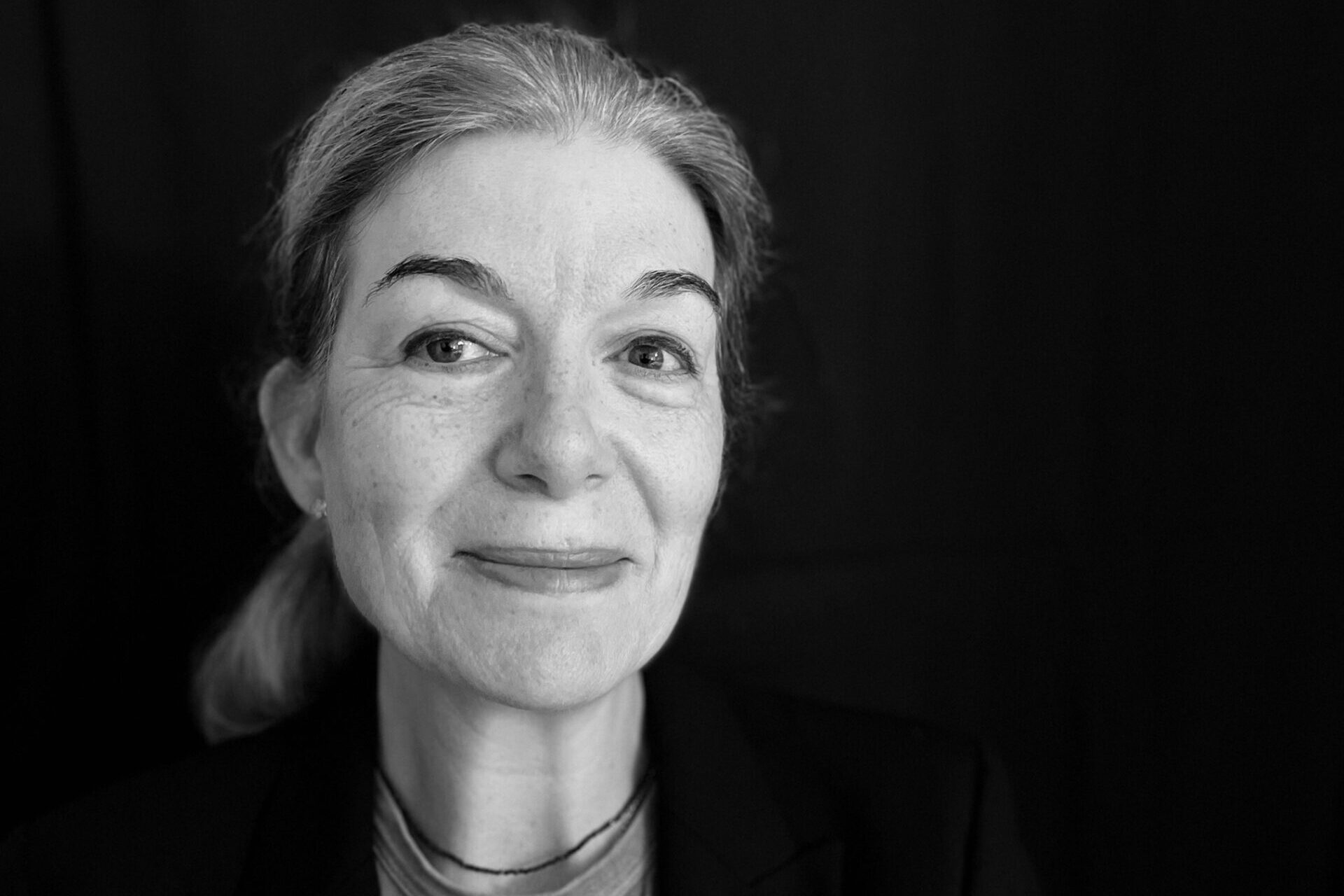 A black-and-white portrait of a person with long hair tied back, wearing a blazer over a shirt. They are looking at the camera with a subtle smile against a plain dark background.