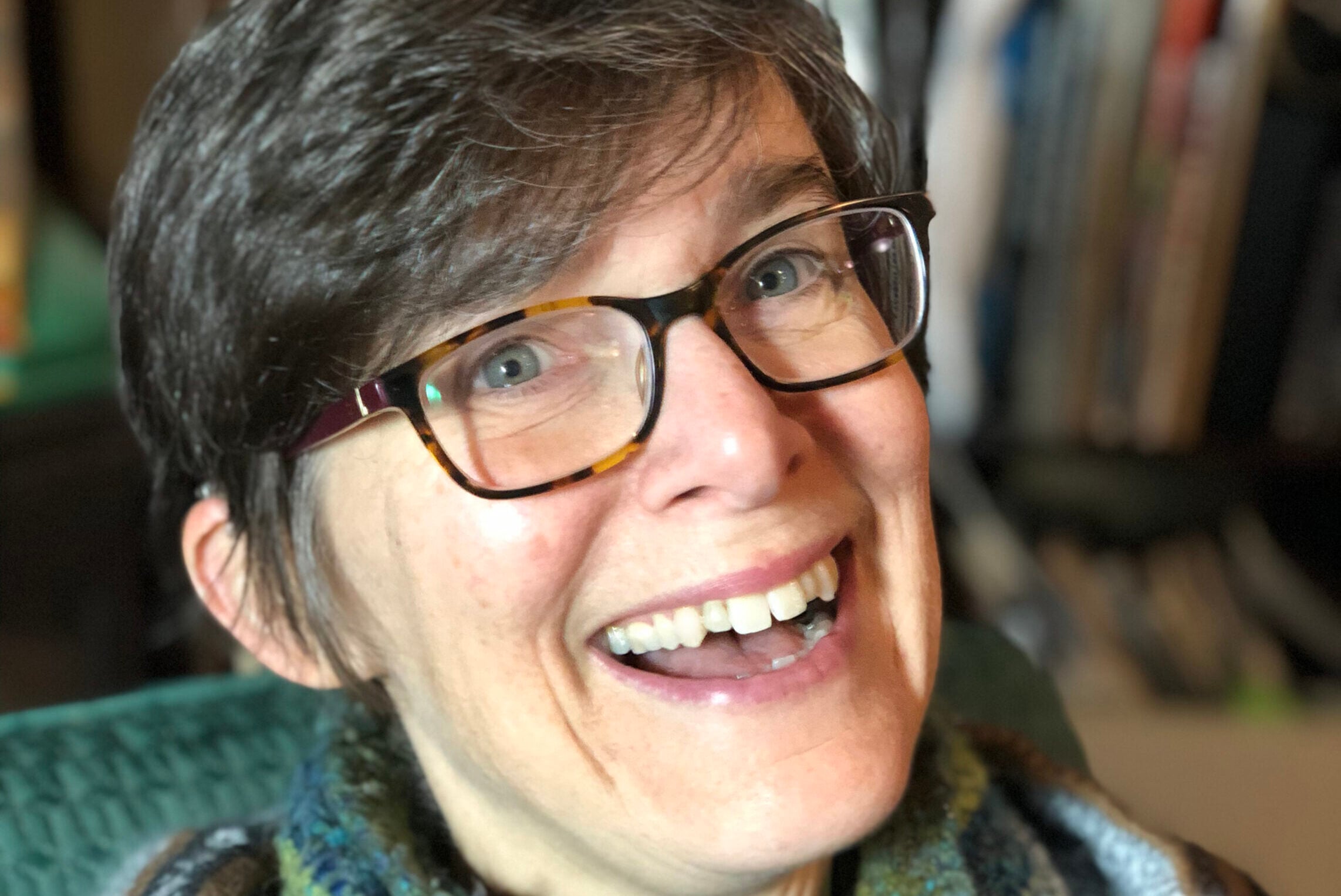 A person with short hair and glasses smiles warmly at the camera. They are wearing a cozy striped sweater and a multicolored scarf, sitting indoors with a bookshelf in the background.
