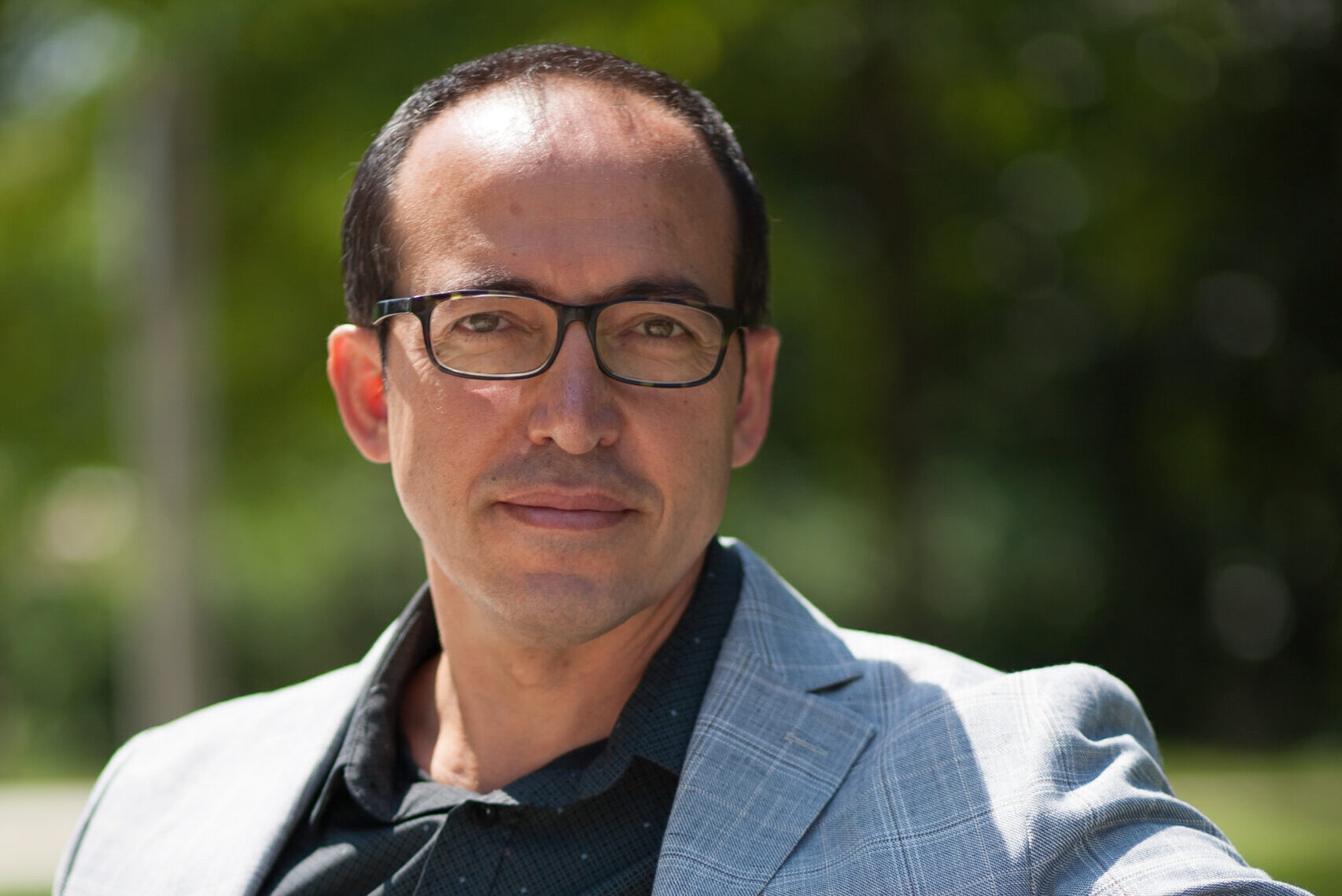 A man wearing glasses and a light gray blazer sits on a bench outdoors. The background is blurred with greenery, and he looks directly at the camera with a neutral expression.