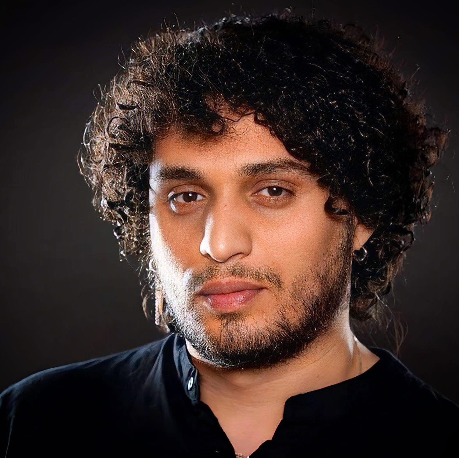 A person with curly hair and a beard is wearing a black shirt and looking directly at the camera against a dark background.
