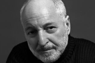 Black and white portrait of an older person with a beard and short hair, wearing a dark ribbed sweater. They are looking directly at the camera with a neutral expression against a plain background.