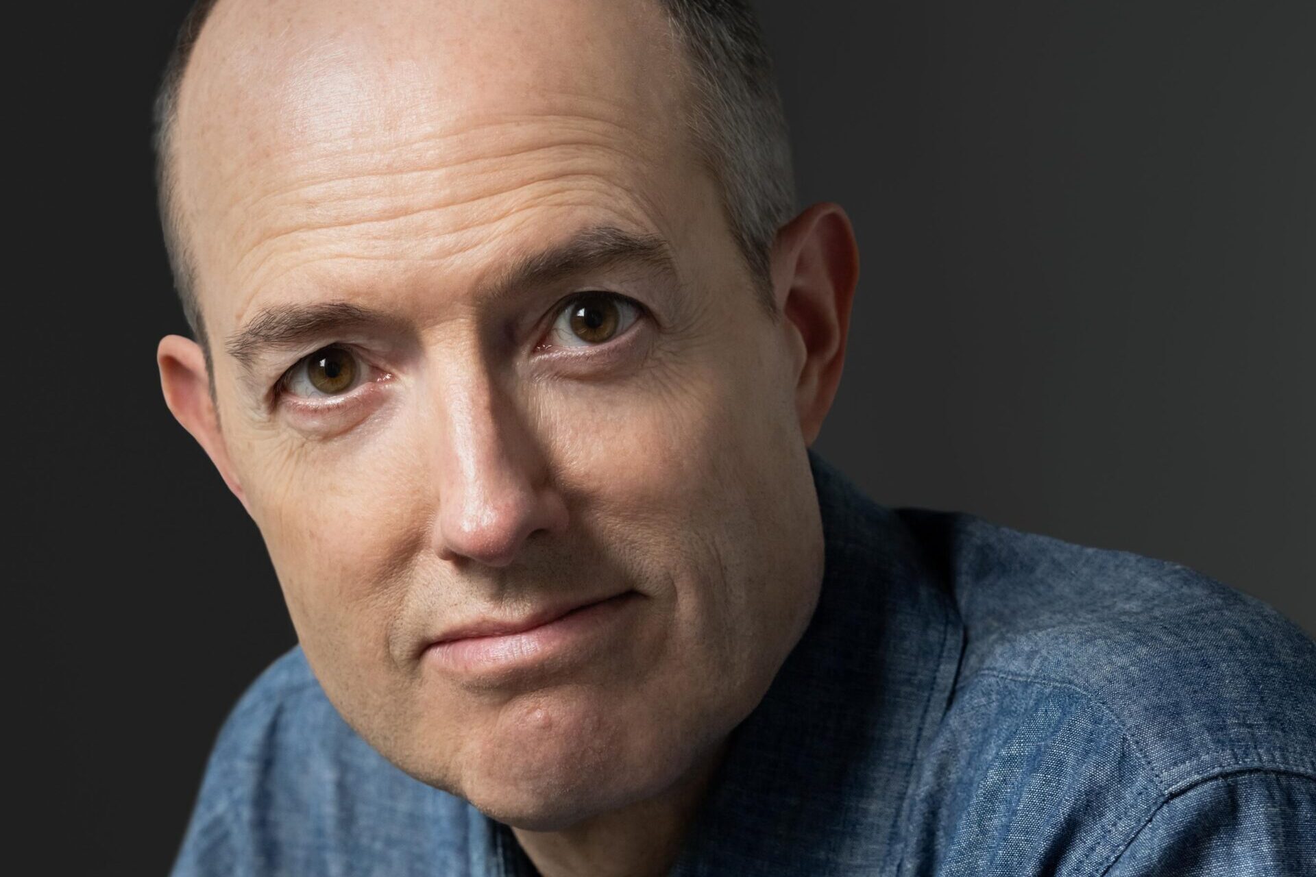 A person with a bald head is wearing a blue button-up shirt and looking at the camera. The background is dark gray, highlighting the persons face.