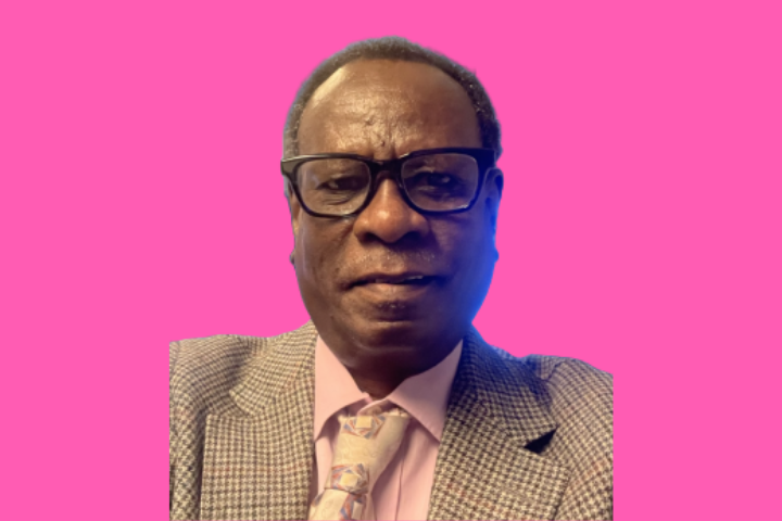 A man wearing glasses, a checked suit jacket, a pink shirt, and a patterned tie is smiling slightly against a bright pink background.