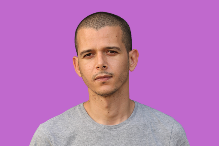A man with short hair and a neutral expression wearing a light gray t-shirt, standing in front of a solid purple background.