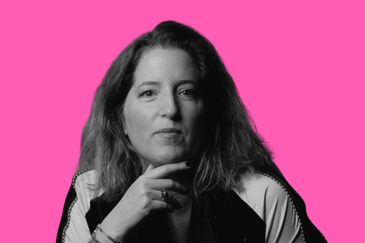 A woman with shoulder-length hair looks thoughtfully at the camera, resting her chin on her hand. She wears a light and dark jacket. The background is solid bright pink.