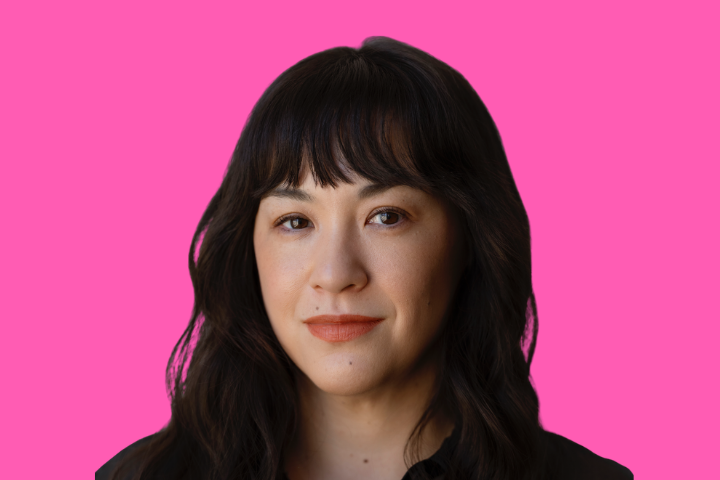 A person with long wavy dark hair and bangs, wearing a dark top, poses in front of a bright pink background.