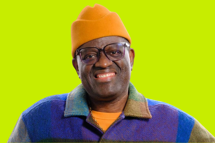 A smiling man wearing glasses, an orange hat, and a colorful jacket stands against a bright green background.