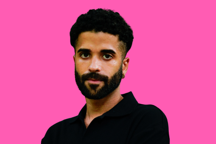 A man with short curly hair and a beard, wearing a black collared shirt, stands in front of a bright pink background, looking directly at the camera with a neutral expression.