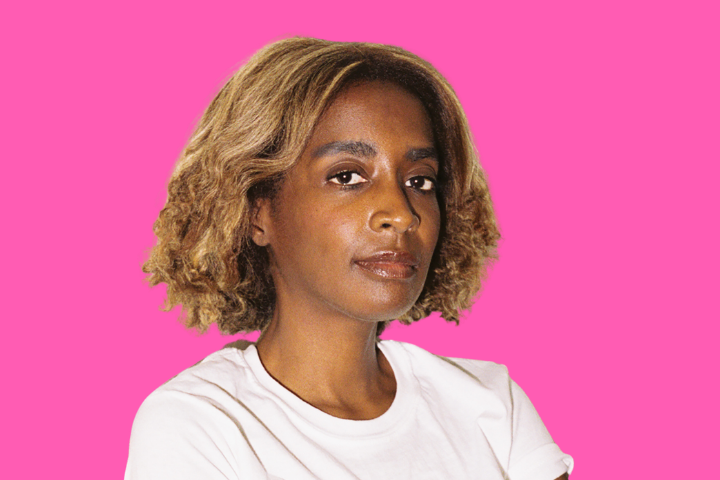 A person with curly, shoulder-length blonde hair and medium skin tone wears a white t-shirt, looking confidently at the camera against a bright pink background.
