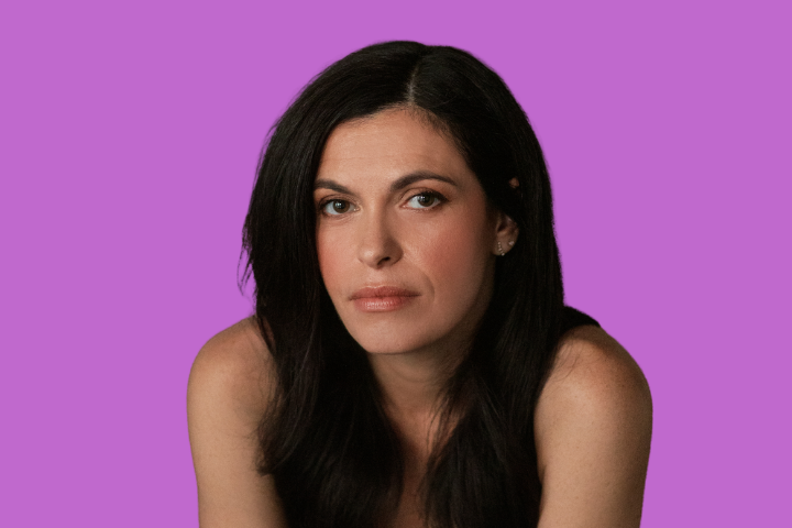 A woman with long dark hair and a neutral expression is looking at the camera. She is wearing a sleeveless top and is posed against a solid purple background.