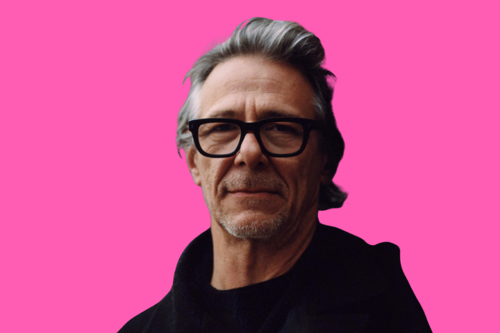 A middle-aged man with gray hair and black glasses, wearing a dark coat, stands against a bright pink background.