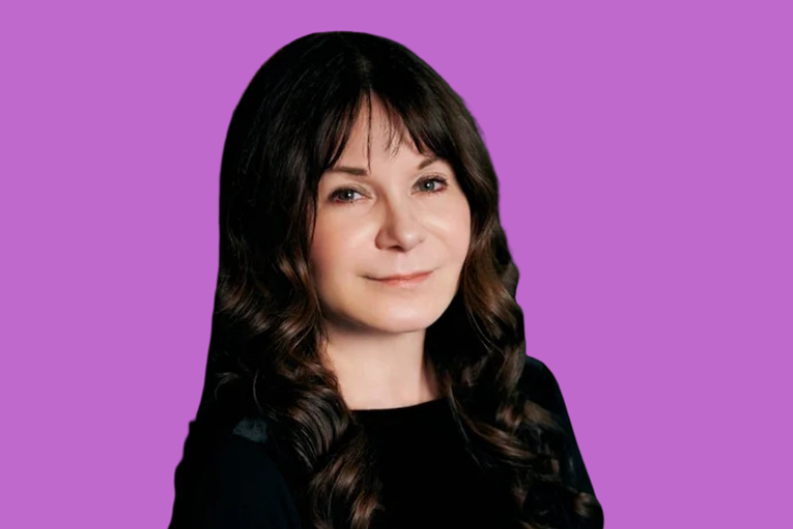 A woman with long, wavy dark hair and bangs, wearing a black top, is smiling softly against a solid purple background.