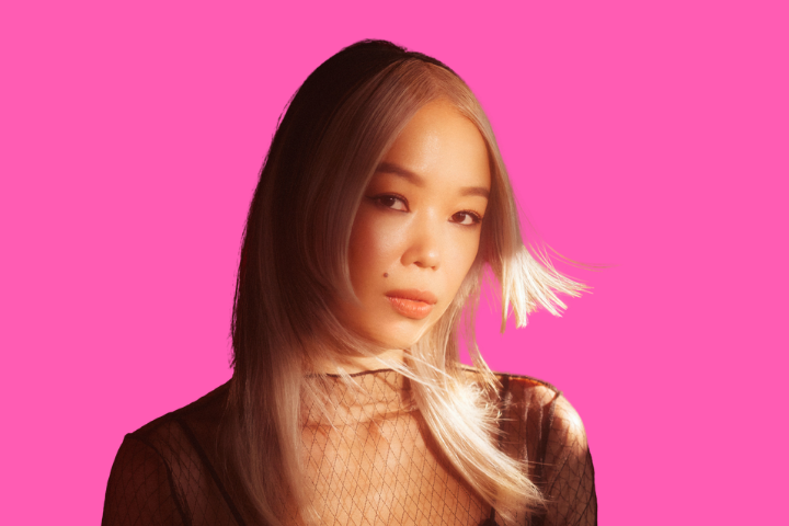 A woman with long, light blond hair and a sheer black top looks calmly at the camera. She is lit by soft, warm light and stands against a bright pink background.