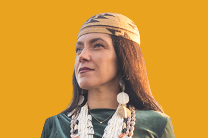 A woman wearing a tan woven headband with geometric patterns, long dark hair, large round earrings, and layered beaded necklaces, stands against a solid mustard yellow background.