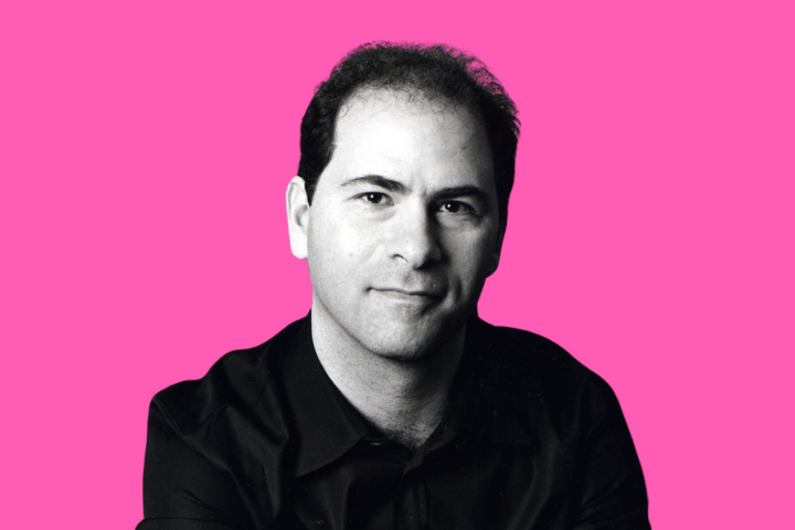 A man with short dark hair wearing a black shirt poses against a solid pink background, looking directly at the camera with a neutral expression.
