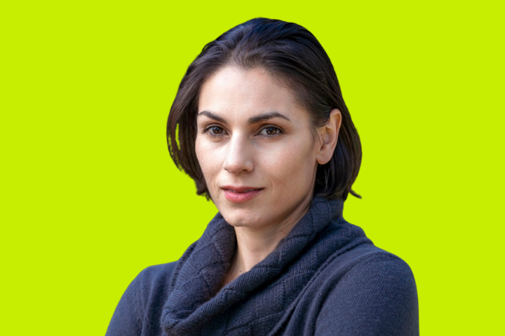 A woman with short, dark hair wearing a dark gray sweater stands against a bright yellow-green background, looking confidently at the camera.