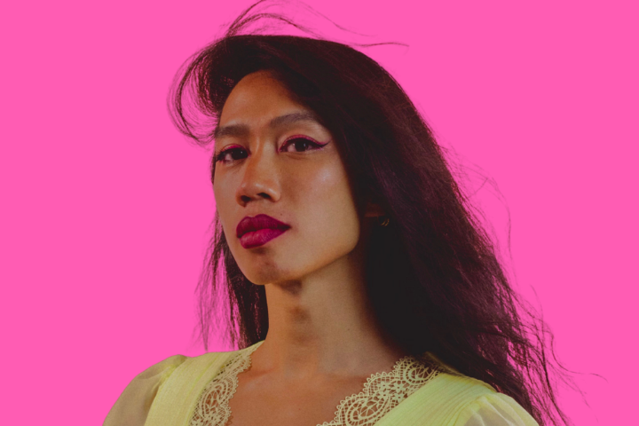 A person with long dark hair and bold pink makeup wears a pale yellow lace-trimmed top, posing against a bright pink background.