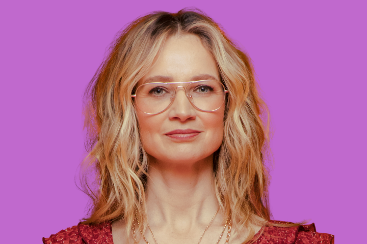 A woman with wavy blonde hair and large glasses is wearing a red patterned top and layered necklaces, standing in front of a solid purple background.