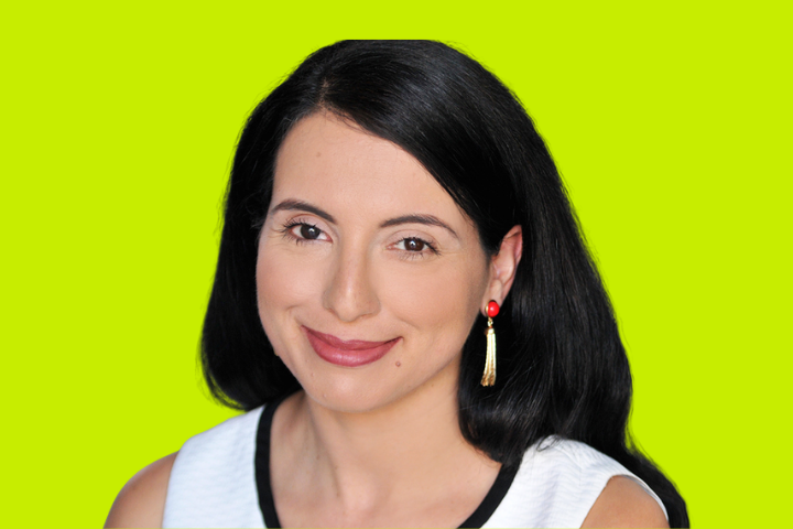 A woman with long black hair, wearing a white top with black trim and gold tassel earrings, smiles at the camera against a bright yellow-green background.