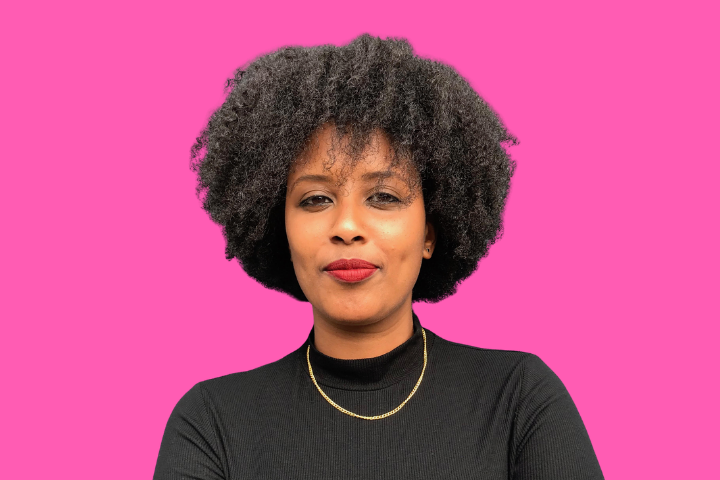 A woman with curly black hair, wearing a black top and a gold necklace, stands in front of a bright pink background, looking confidently at the camera.