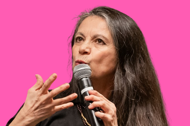 A woman with long dark hair speaks into a microphone, gesturing with her hand. She stands against a bright pink background and wears a black top.