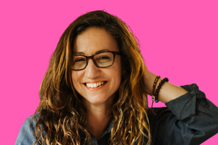 A smiling woman with long wavy hair and glasses, wearing a dark shirt and beaded bracelets, stands against a bright pink background, touching her hair with one hand.