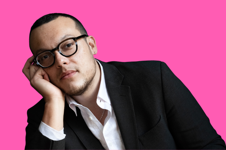 A person with short dark hair, glasses, and a trimmed beard rests their head on their hand, wearing a black suit jacket and white shirt, against a bright pink background.