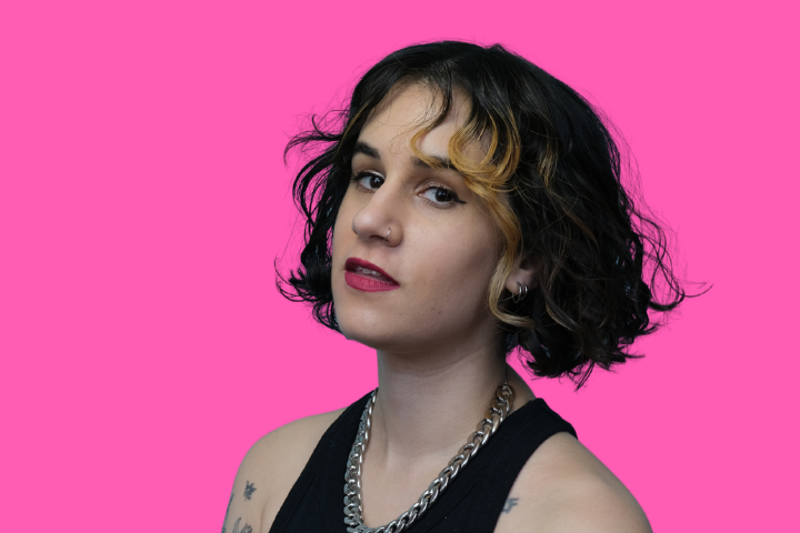 A person with short, wavy dark hair with blonde highlights and a side fringe, wearing a sleeveless black top and a chunky silver chain necklace, poses against a bright pink background.