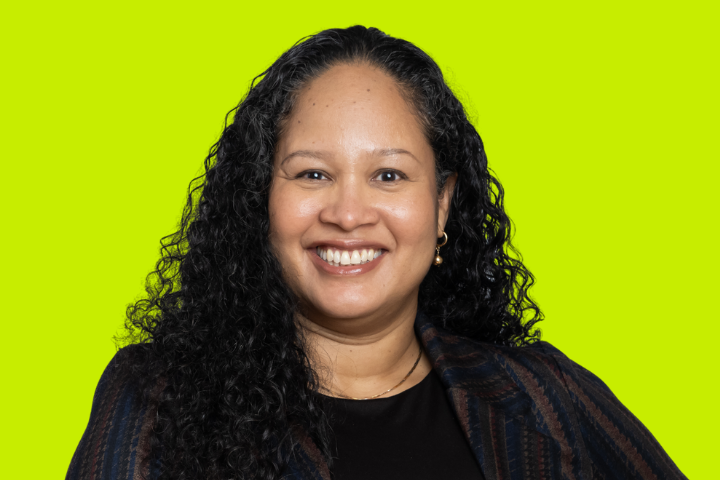 A woman with long, curly black hair smiles at the camera. She is wearing a dark, striped blazer over a black top, with a bright yellow-green background behind her.