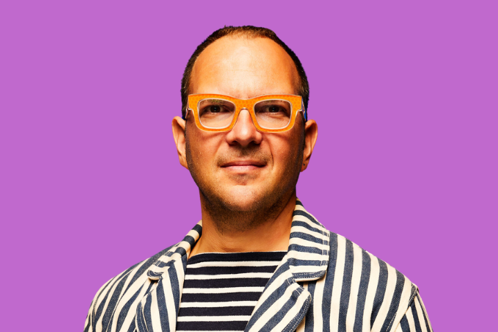 A person wearing orange glasses and a black-and-white striped jacket over a striped shirt smiles slightly in front of a solid purple background.