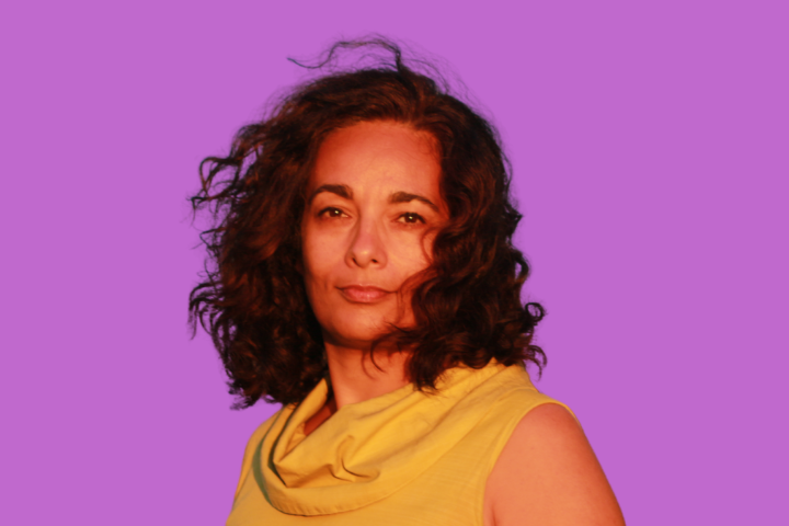 A person with curly brown hair wearing a sleeveless yellow top looks confidently at the camera against a solid purple background.