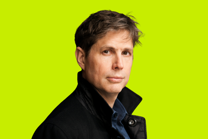 A man with short brown hair wearing a dark jacket looks at the camera against a bright lime green background.