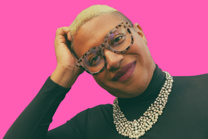 A person with short blond hair, tortoiseshell glasses, and a black top with a layered pearl necklace smiles while resting their head on their hand. The background is bright pink.