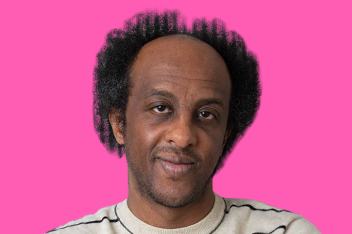 A man with a receding hairline and curly hair looks at the camera with a neutral expression. He is wearing a beige sweater with black stripes. The background is bright pink.