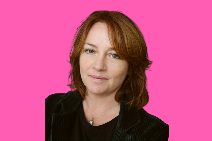 A woman with shoulder-length auburn hair wearing a black top and jacket, looking at the camera with a neutral expression, posed against a solid bright pink background.