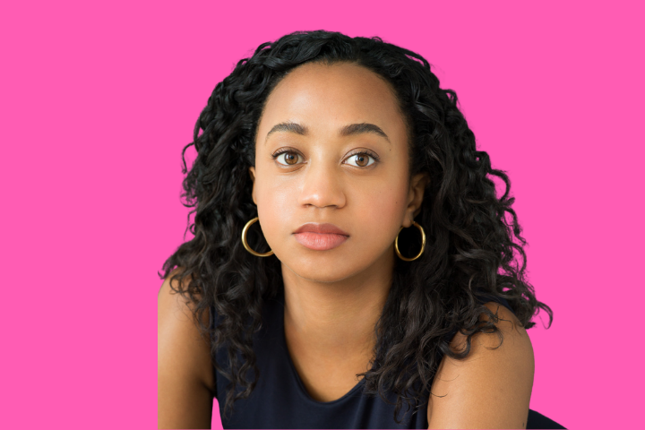 A young woman with curly dark hair and gold hoop earrings looks at the camera with a neutral expression, set against a solid pink background. She is wearing a sleeveless dark top.