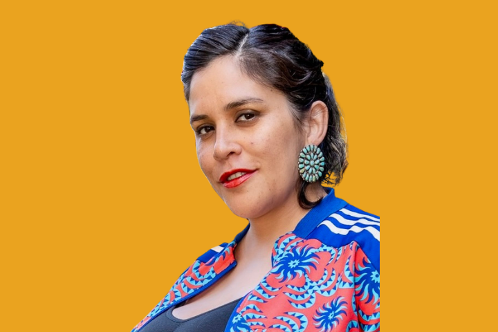 A woman with dark hair styled back wears large green earrings, a colorful patterned jacket with blue and orange designs, and a black top, posing against a solid orange background.