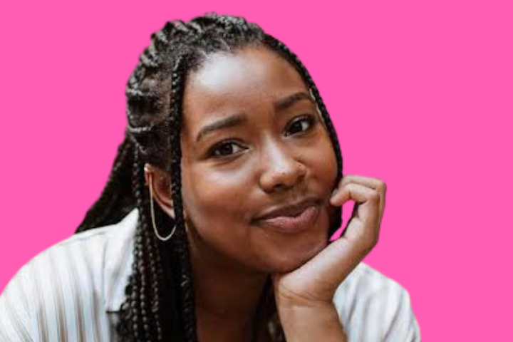 A woman with long braided hair, wearing hoop earrings and a striped shirt, rests her chin on her hand and smiles gently against a bright pink background.