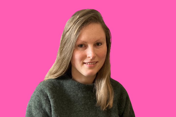 A person with long, straight, light brown hair and a dark sweater is smiling slightly, sitting in front of a bright pink background.