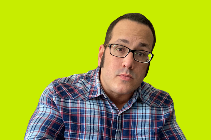 A person wearing glasses and a blue plaid shirt looks directly at the camera, against a bright yellow-green background.