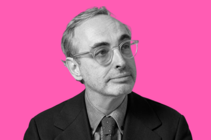 A man with short, light hair and glasses wearing a suit and tie looks slightly upward. The photo is black and white, and the background is bright pink.