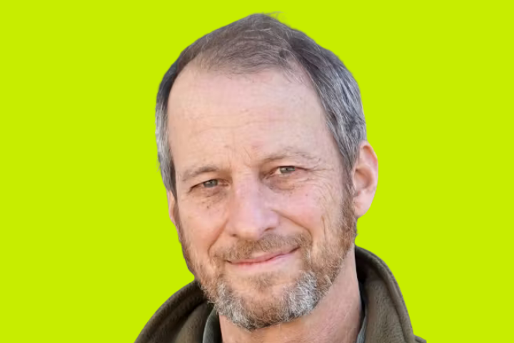 A middle-aged man with short, graying hair and a beard smiles softly. He is wearing an olive green jacket, and the background is a solid bright yellow-green.