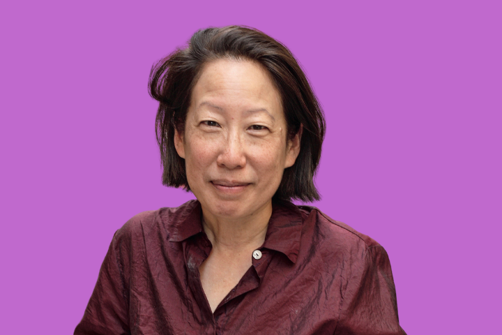 A person with short dark hair wearing a maroon button-up shirt, smiling slightly, is posed in front of a plain purple background.
