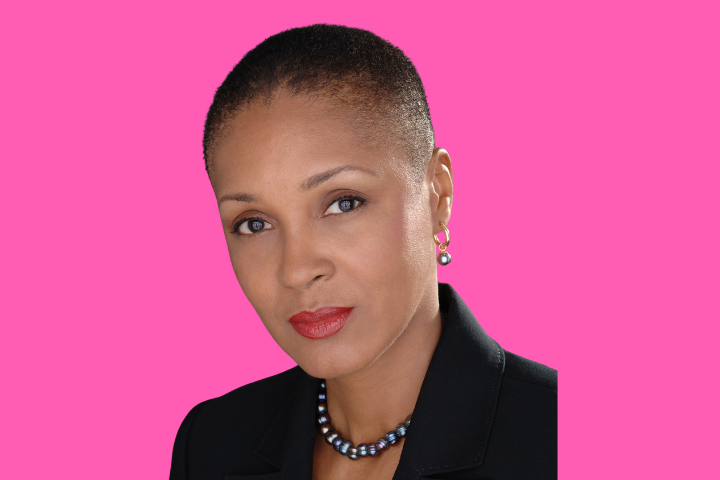 A woman with short hair, wearing a black blazer, pearl earrings, and a beaded necklace, poses against a bright pink background.