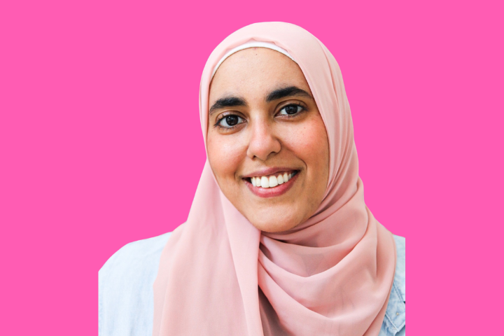 A woman wearing a light pink hijab and light-colored top smiles at the camera, posed against a bright pink background.