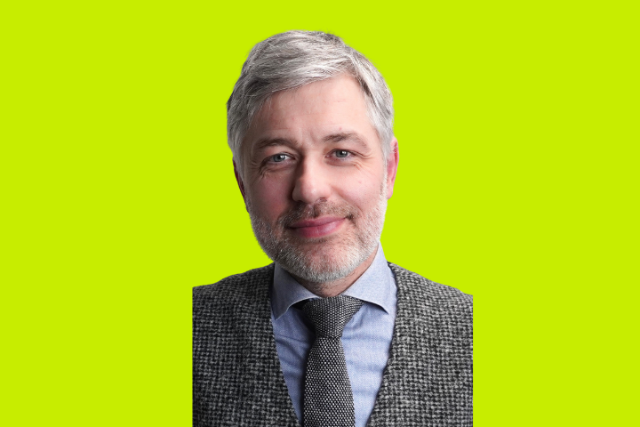 A middle-aged man with short gray hair and a beard, wearing a gray patterned suit, blue shirt, and tie, stands in front of a bright yellow-green background, smiling gently.